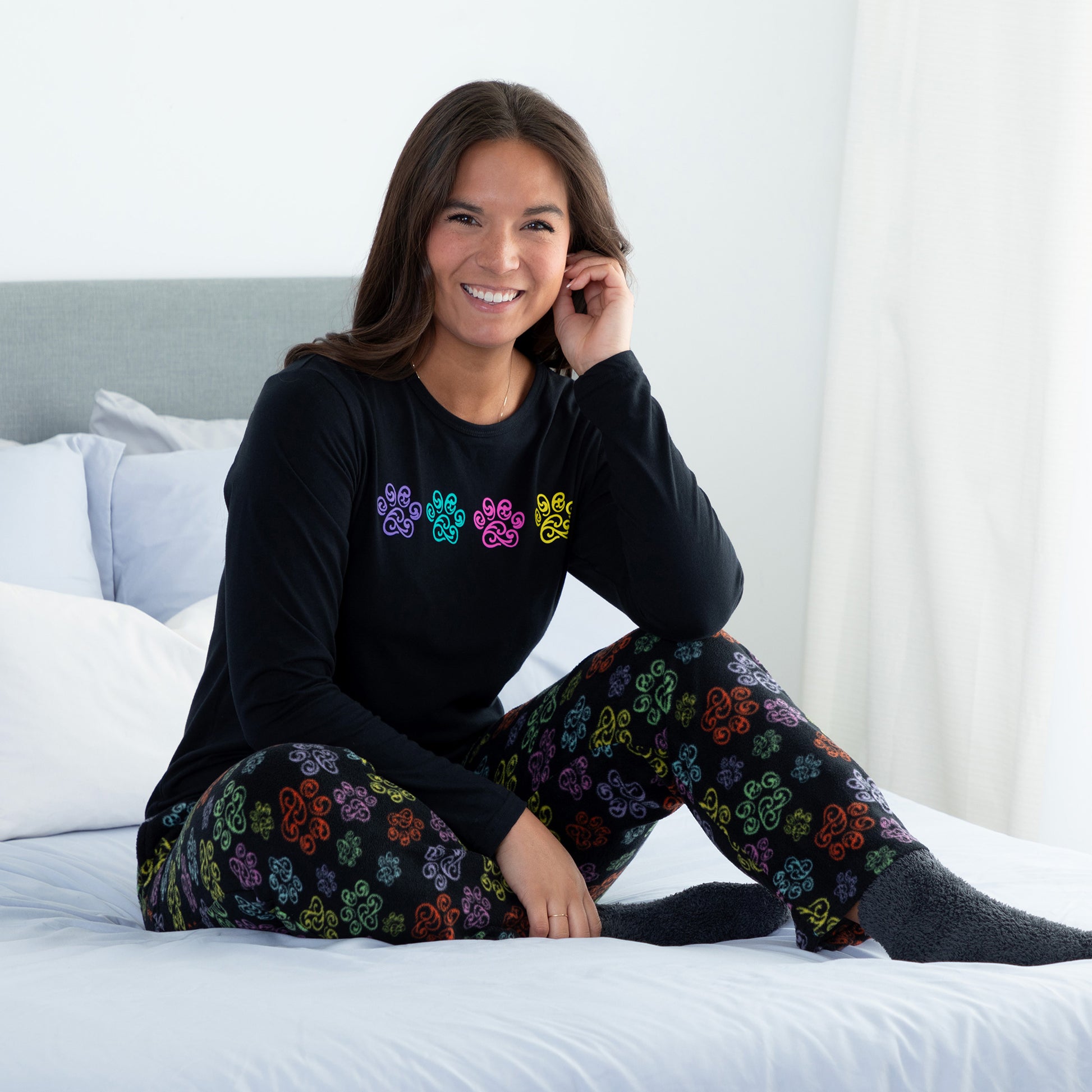 A woman sitting on a bed in a bedroom setting wearing a black long-sleeve top with swirling multicolored paw print and with design swirling multicolored paw print pants