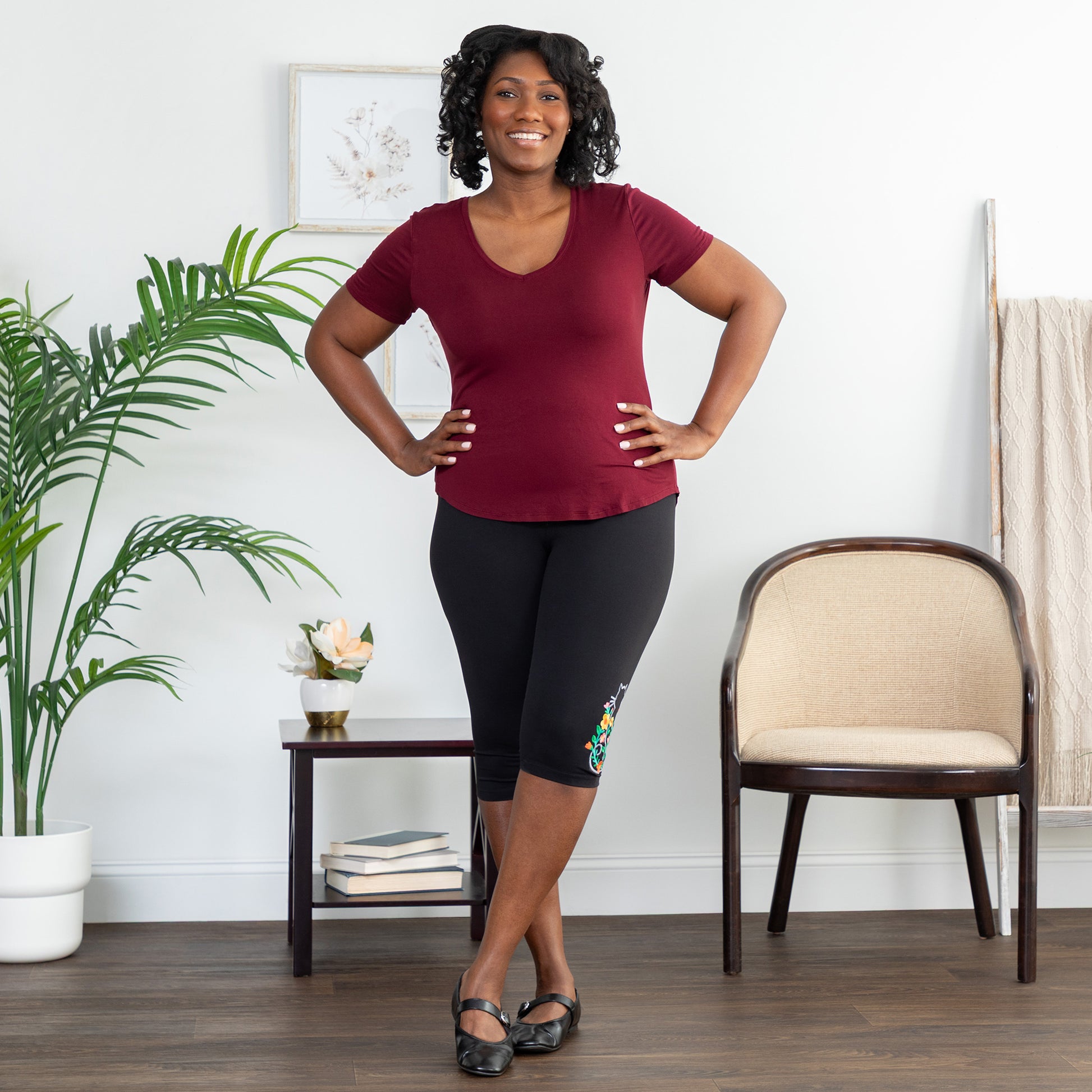 Woman in maroon top and black capri leggings standing in a room with a plant and chair.