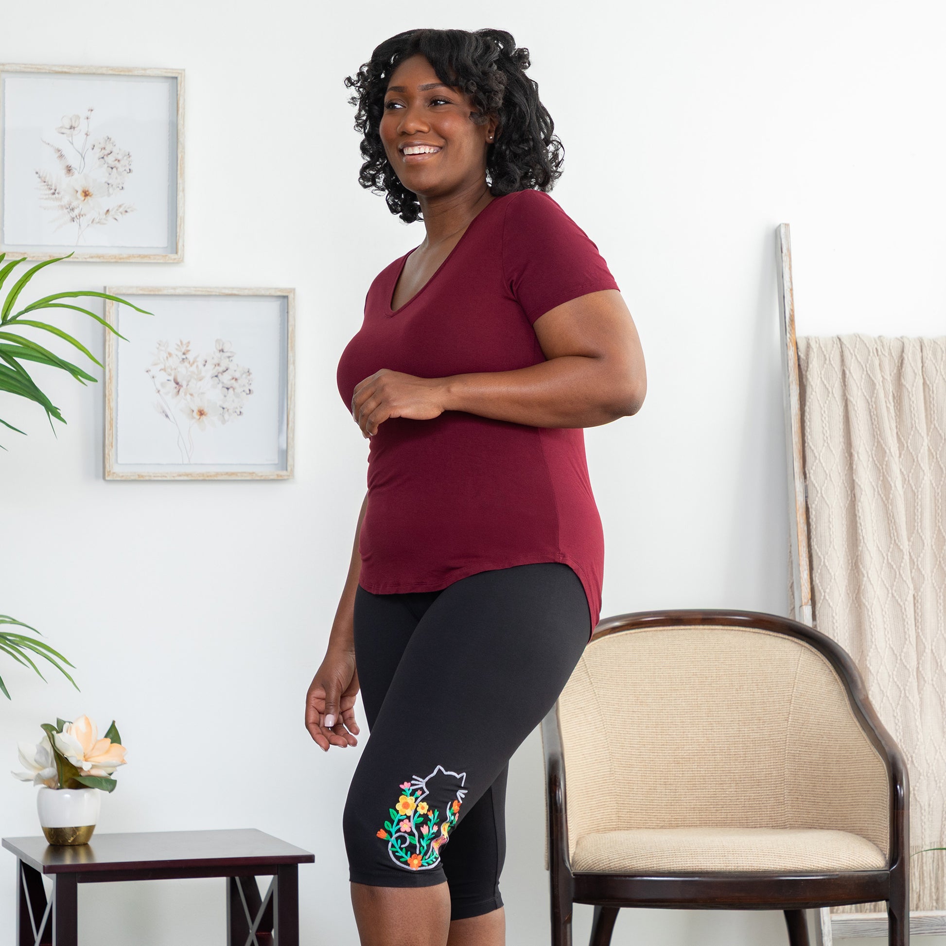 Woman wearing a maroon shirt and black legging capris with a cat and floral design in a living room.