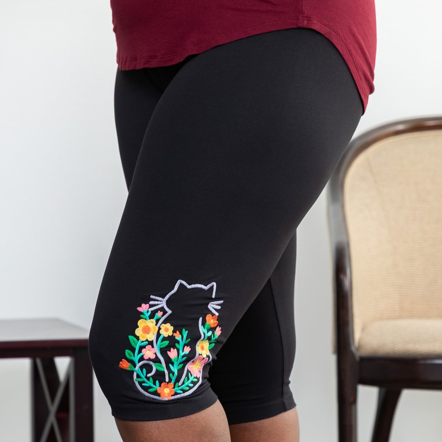 Black leggings with a colorful floral cat design worn by a person, with a chair in the background.