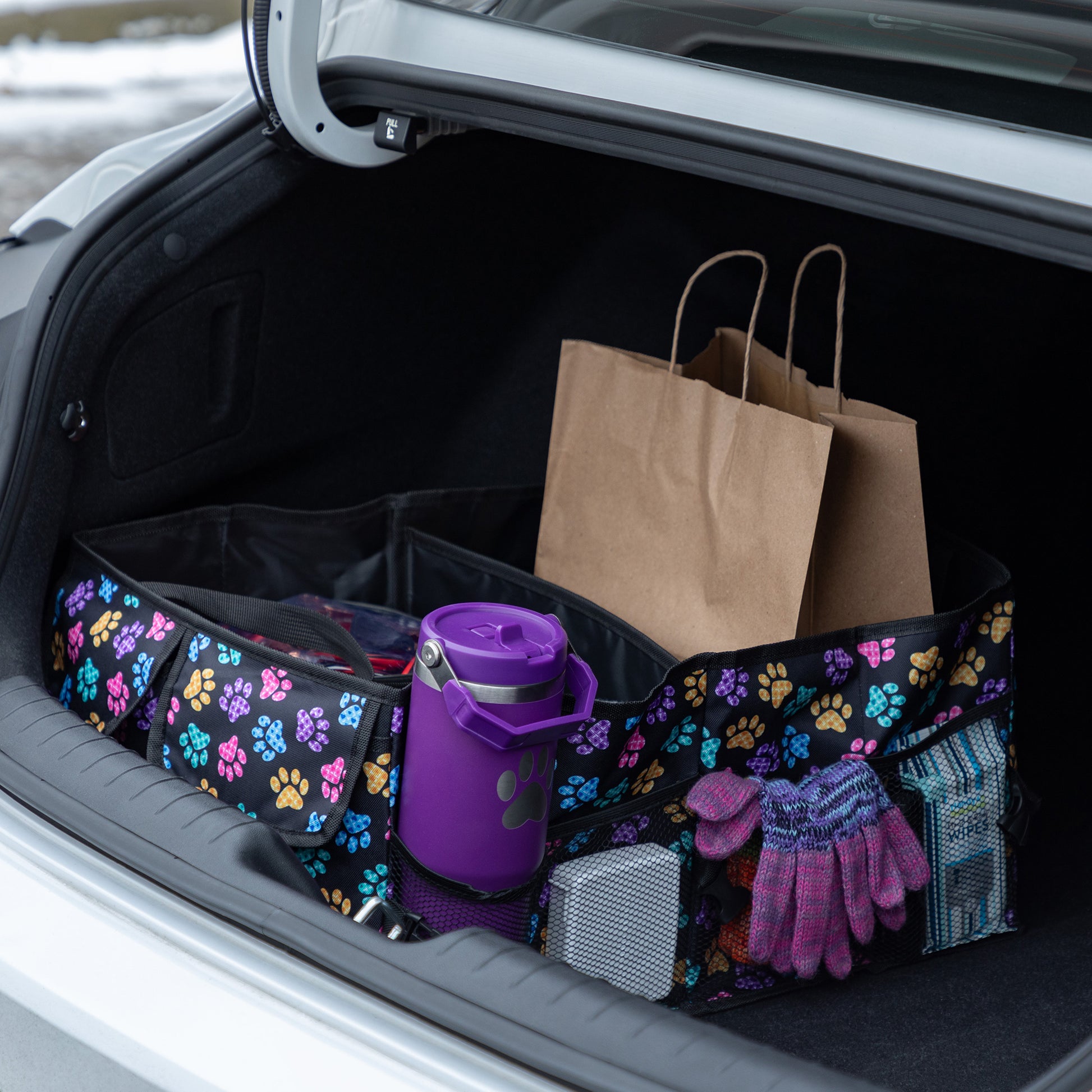 A purple paw printed car trunk organizer, in a car trunk, filled with grocery bags, water bottle, gloves, and other items.