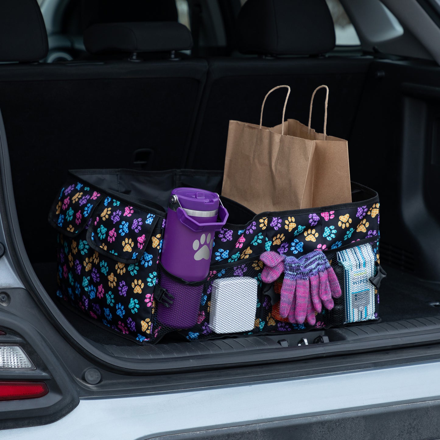 A purple paw printed car trunk organizer, in a car trunk, filled with grocery bags, water bottle, gloves, and other items.