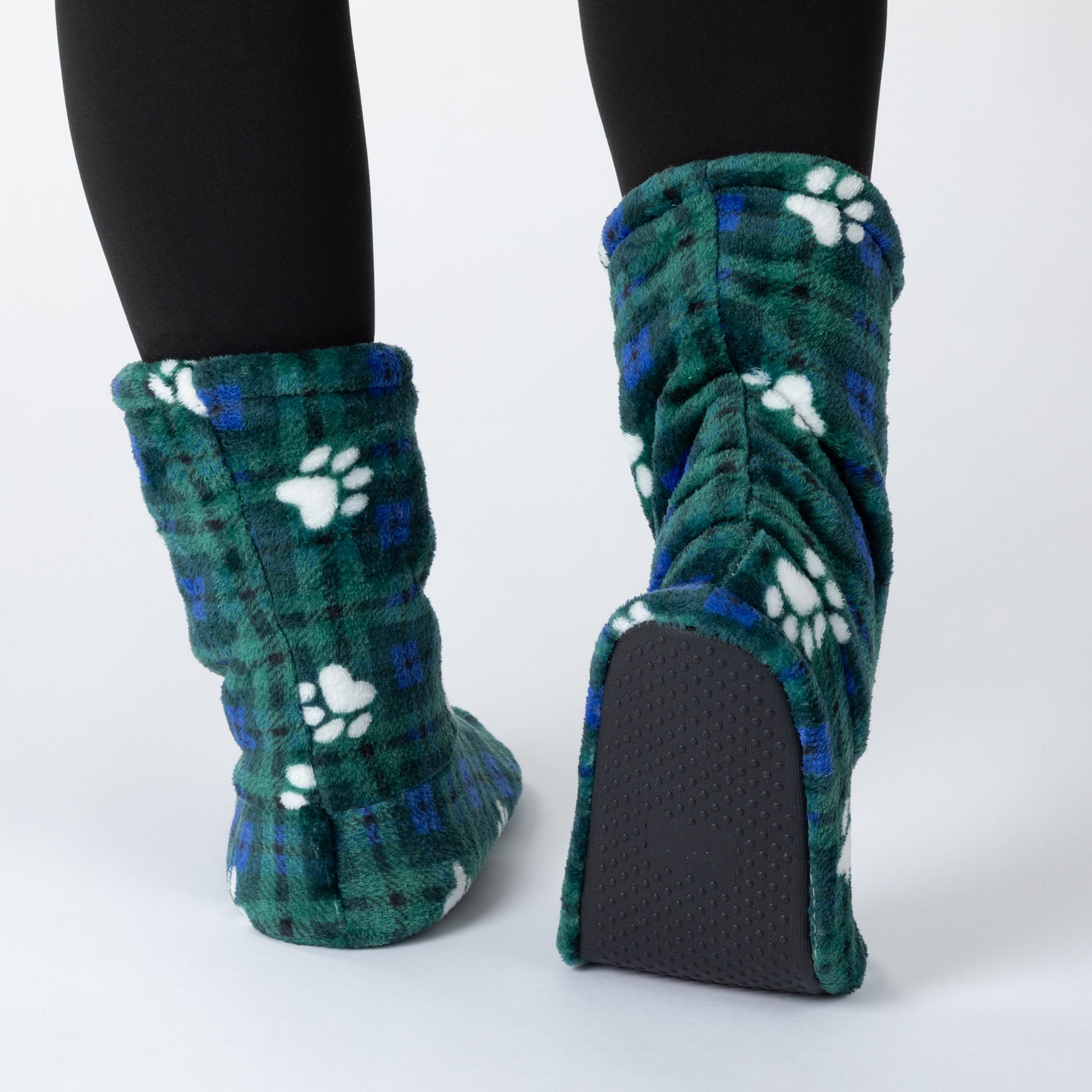 A pair of plush fleece slipper boots with a green and blue plaid pattern and white paw prints, shown on feet wearing black tights against a light gray background.