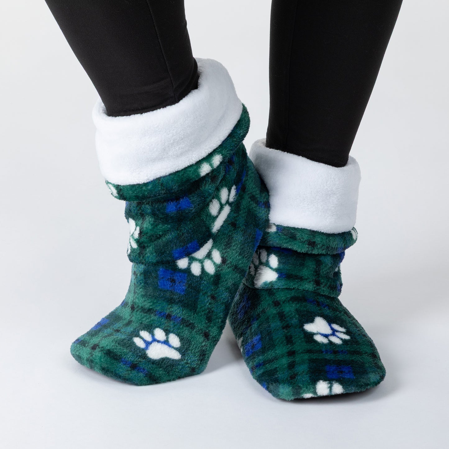 A pair of plush fleece slipper socks with a green and blue plaid pattern, white paw prints, and a white fleece cuff, shown on a person's feet wearing black leggings against a light gray background.