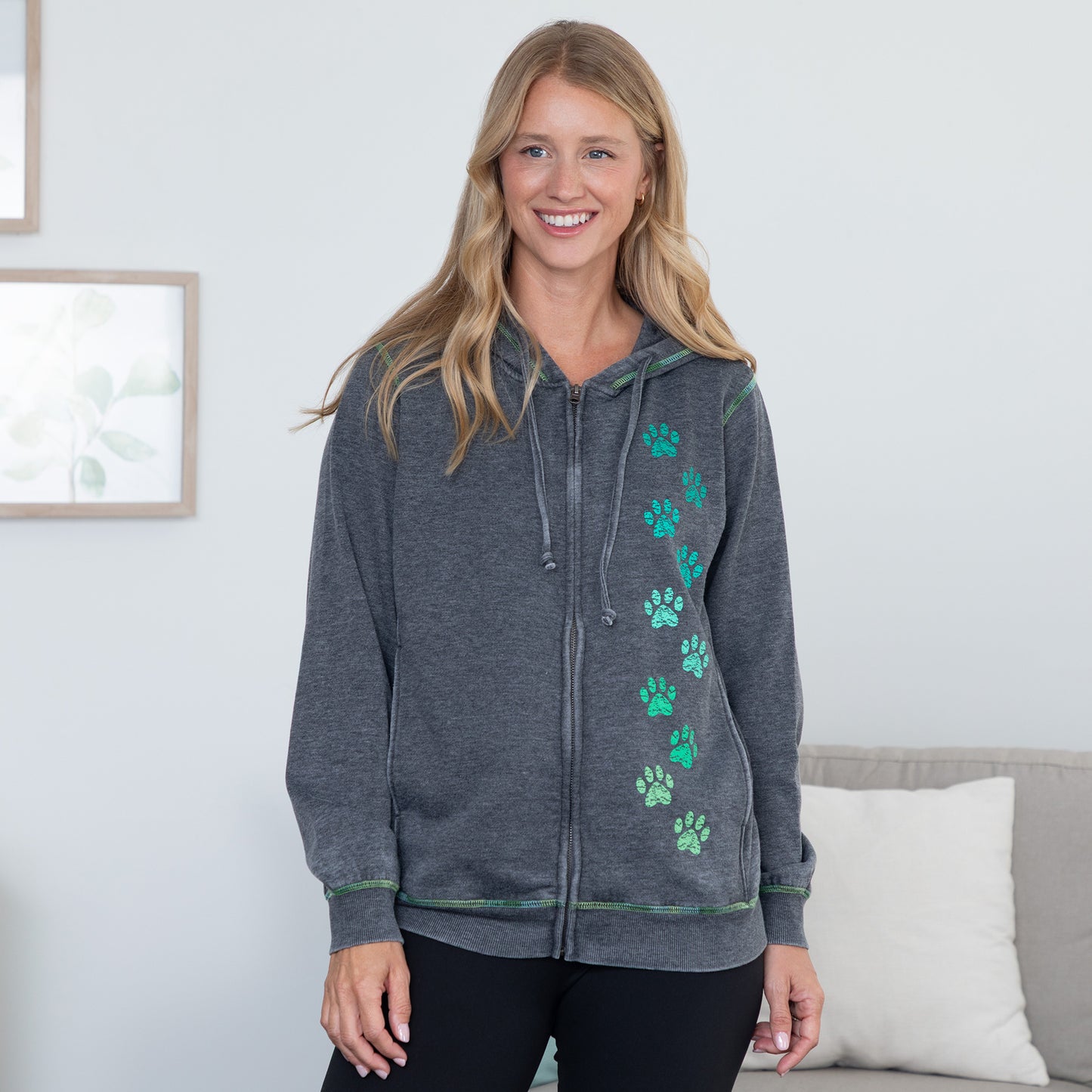 A woman wearing a dark gray zip-up hoodie with green paw prints down the front, standing in a brightly lit room with a sofa and framed wall art in the background.