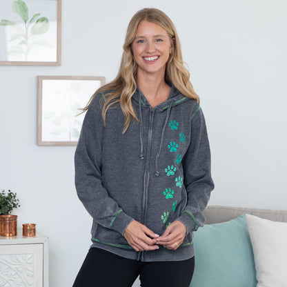 A smiling blonde woman stands indoors wearing a dark heather gray zip-up hoodie with green paw print graphics down the front and green trim details. She is wearing black pants, with a couch, pillows, and framed wall art visible in the background.
