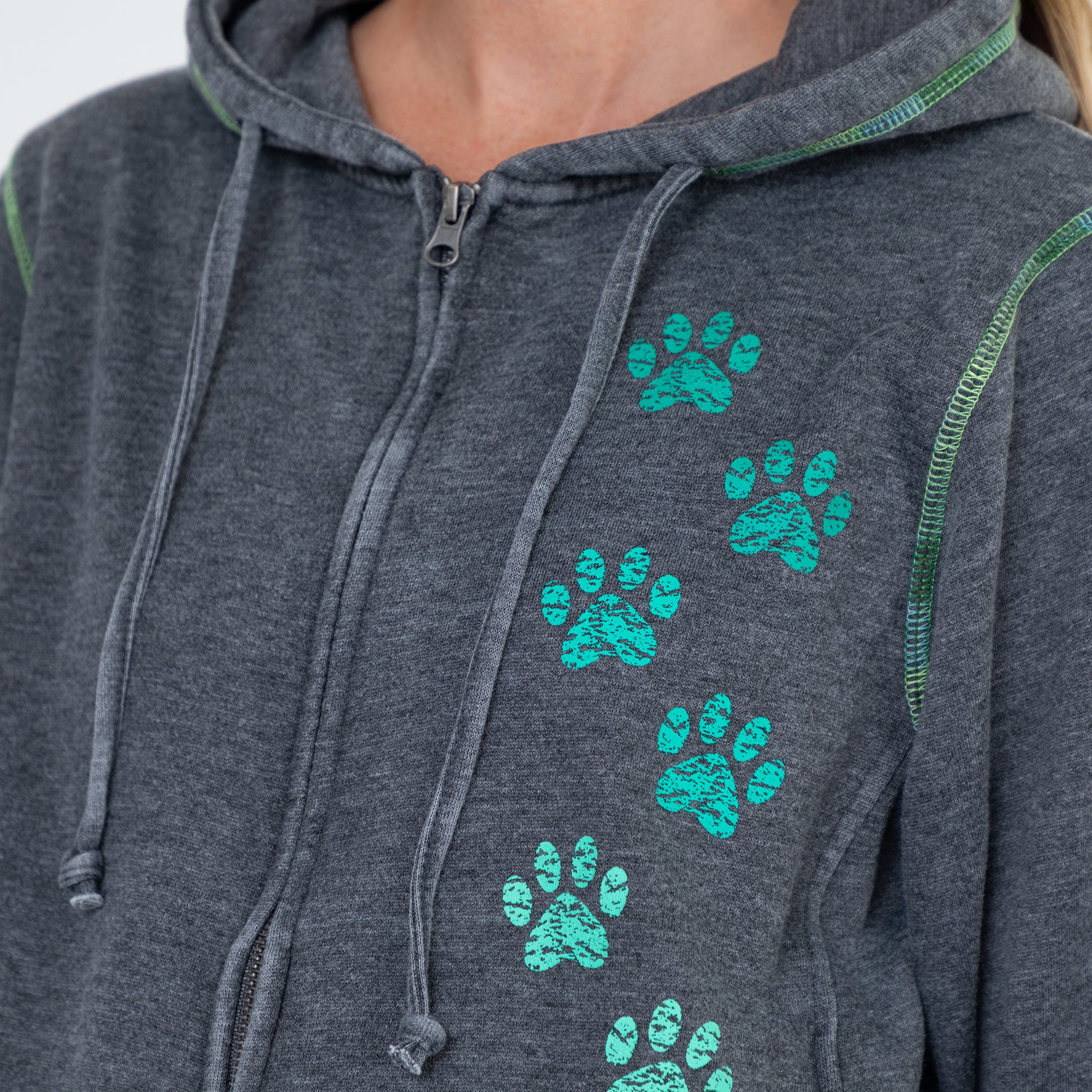 Close-up of a dark heather gray zip-up hoodie worn by a model, featuring a vertical pattern of bright teal paw prints on the front and green contrast stitching.