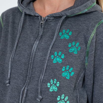 Close-up of a dark heather gray zip-up hoodie worn by a model, featuring a vertical pattern of bright teal paw prints on the front and green contrast stitching.
