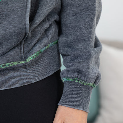 Close-up of a person wearing a heather gray ribbed hoodie with green contrast stitching along the cuffs and hem, paired with black bottoms.