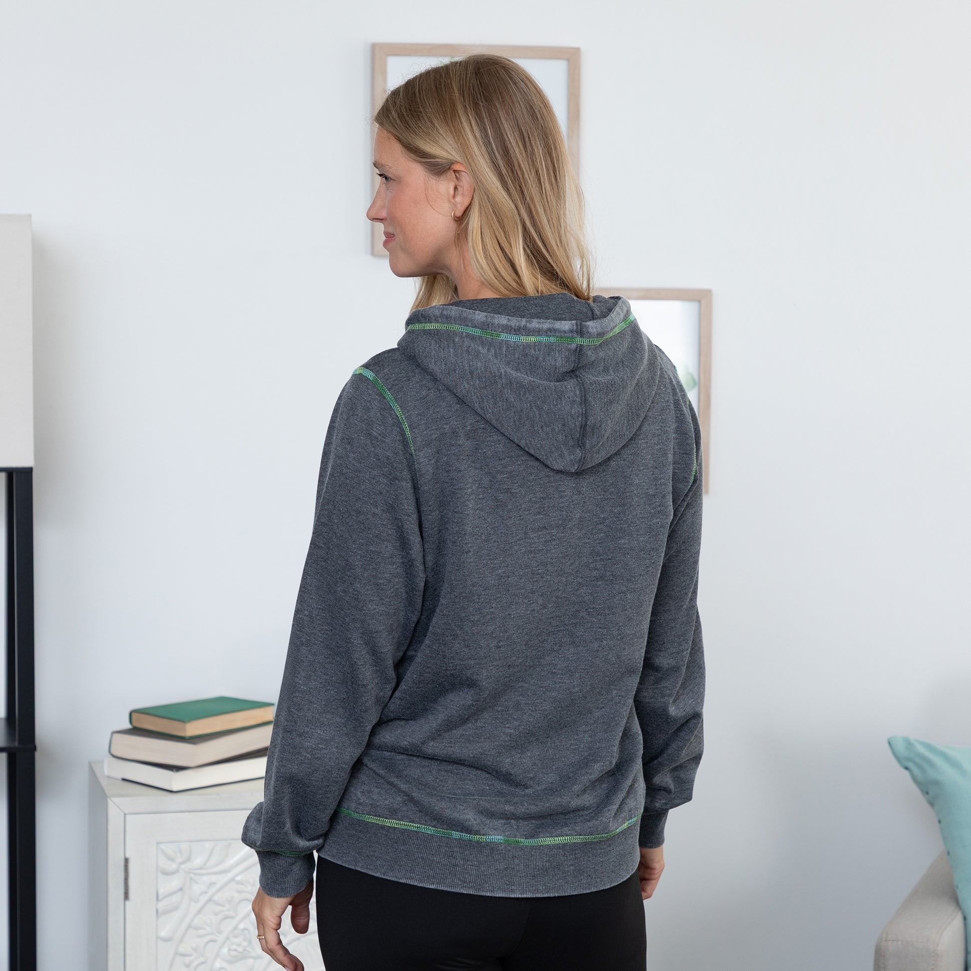 A woman shown from the back wearing a dark heather gray hoodie with thin green contrast stitching, standing in a brightly lit room.