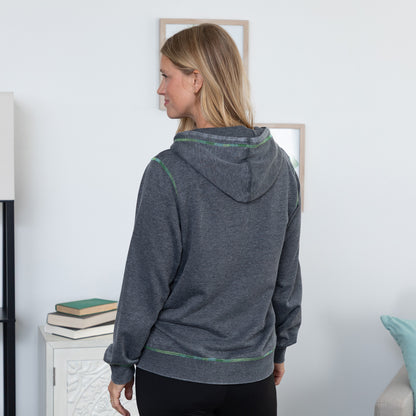 A woman shown from the back wearing a dark heather gray hoodie with thin green contrast stitching, standing in a brightly lit room.