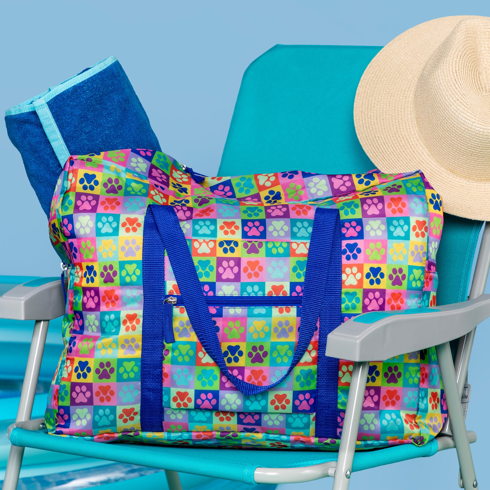 A colorful paw print checkered duffel bag with blue handles and zippers resting on a beach chair against a blue background. Coming out of the duffel bag is a large beach towel.  