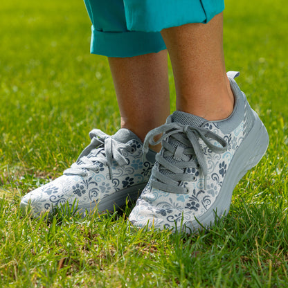 A close-up of a person wearing light gray and white floral-patterned sneakers with gray laces, standing on green grass. The person is also wearing teal cropped pants.