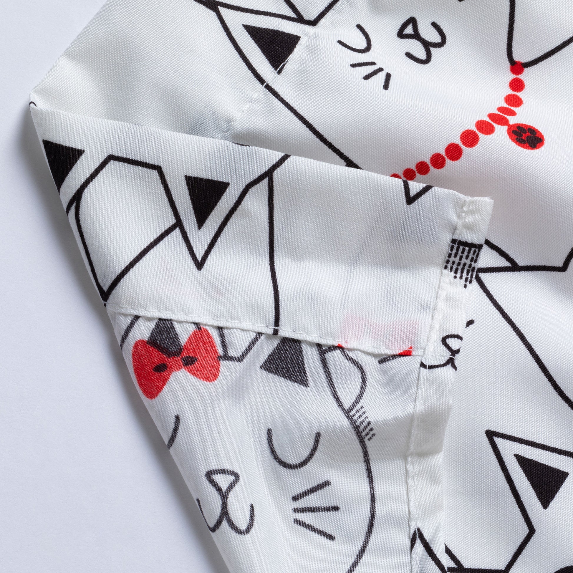 Close-up of a white fabric product printed with black cat face outlines and red bow tie and necklace details, showing the fabric texture and stitching.