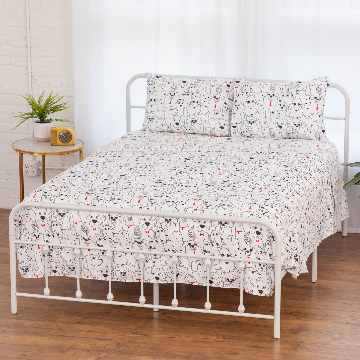 A bed dressed in a white sheet and pillowcase set featuring a black line-art cat pattern with small red accents, styled in a bright bedroom with a white metal bed frame, wood floor, and a small side table.