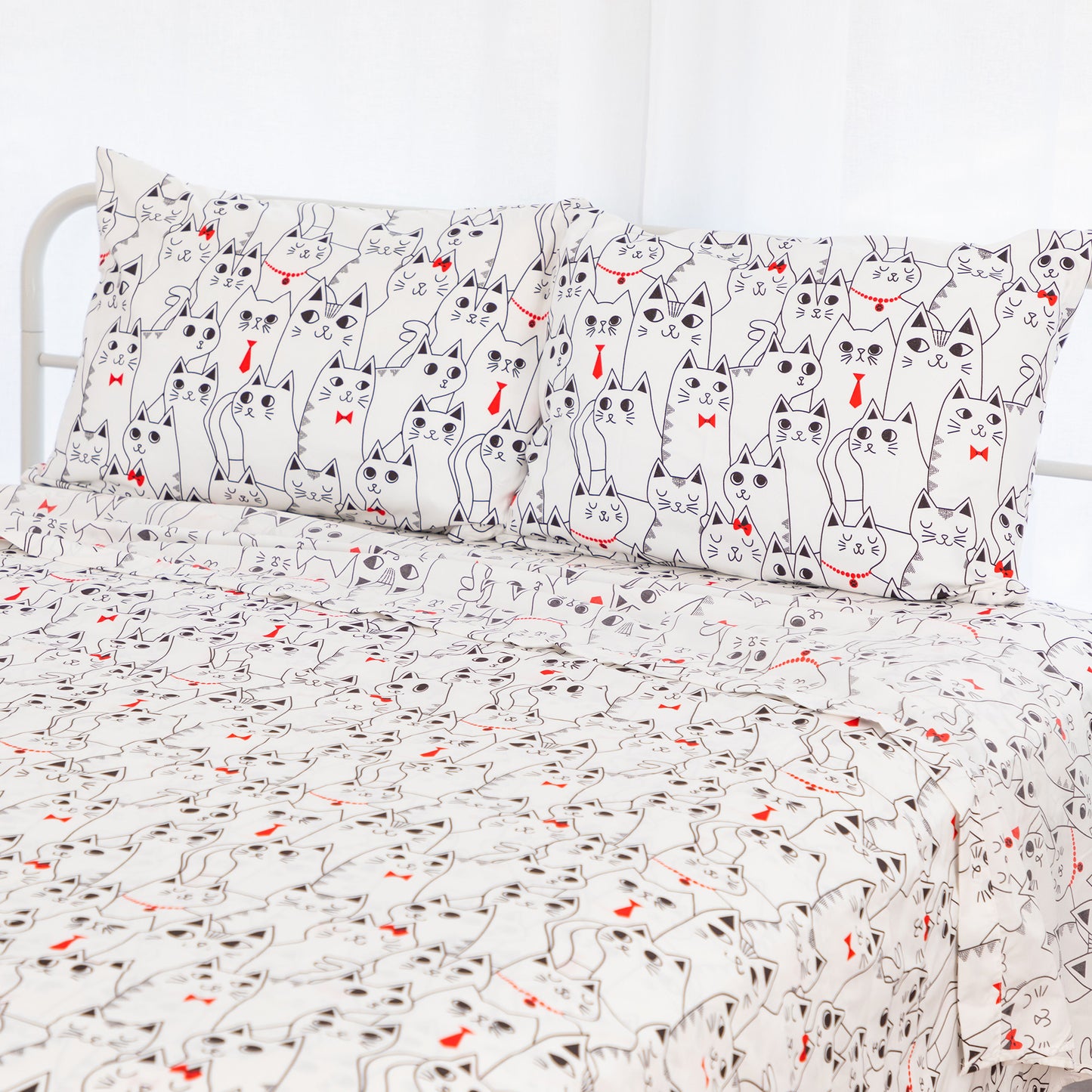 A bed dressed in a white bedding set featuring a repeating cartoon cat print with black outlines and small red bow ties or heart accents, shown with two matching pillowcases.
