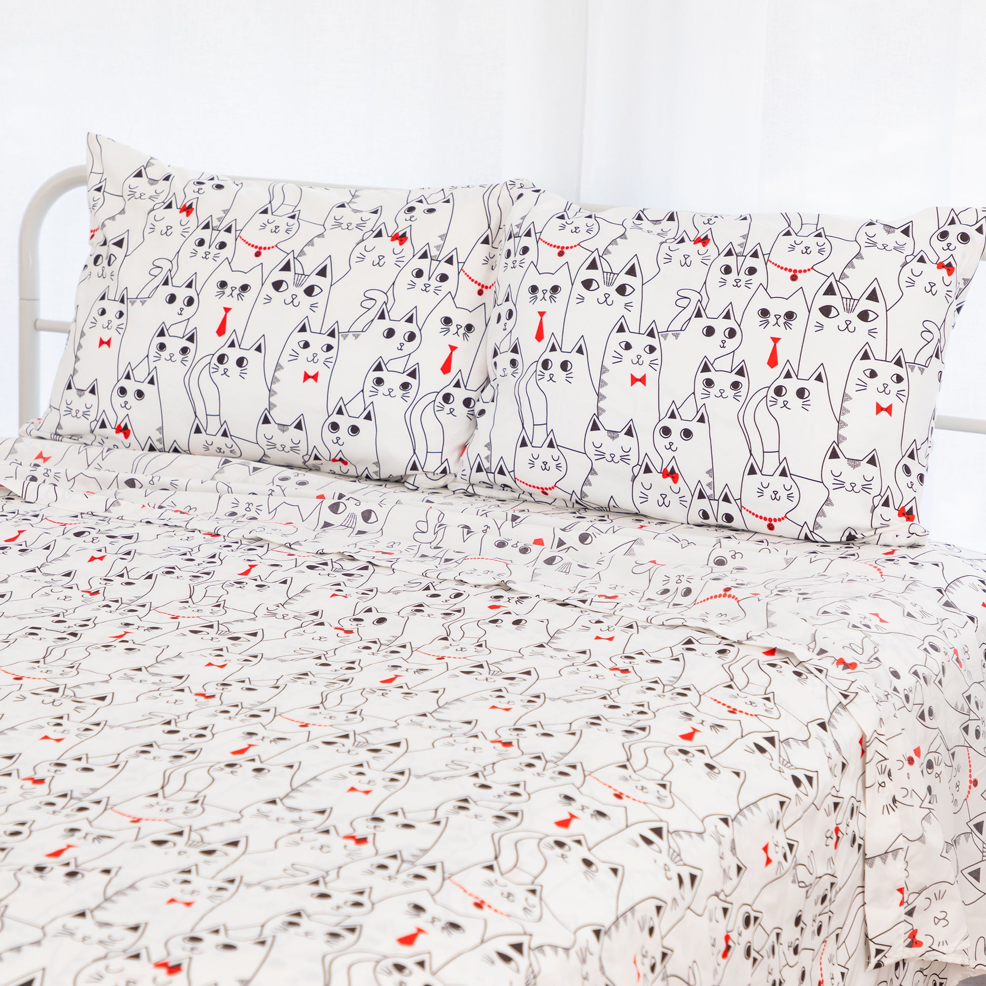 A bed dressed in a white bedding set featuring a repeating cartoon cat print with black outlines and small red bow ties or heart accents, shown with two matching pillowcases.