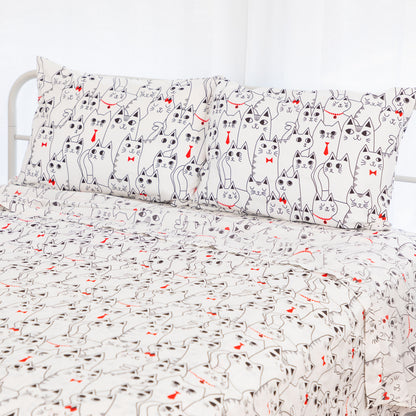 A bed dressed in a white bedding set featuring a repeating cartoon cat print with black outlines and small red bow ties or heart accents, shown with two matching pillowcases.