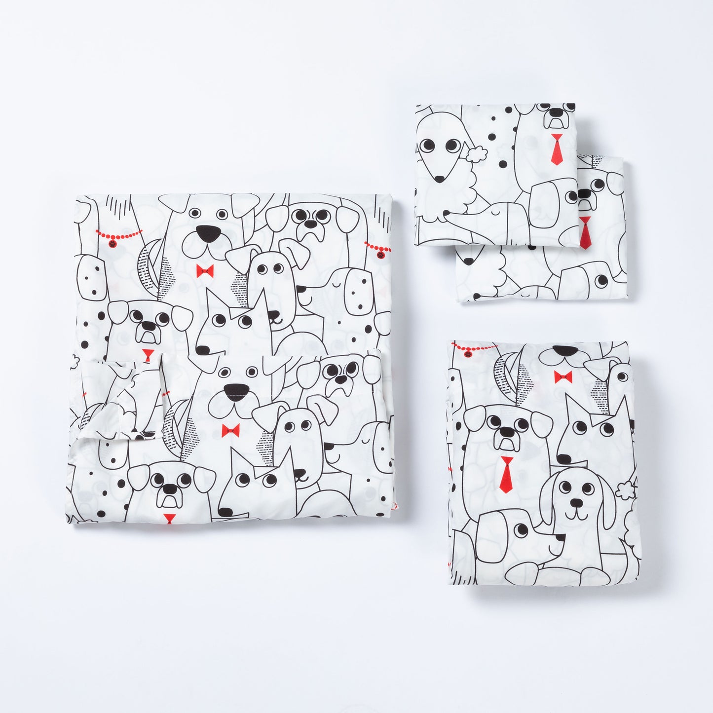 A set of folded sheets, including a larger square piece and three smaller rectangular pieces, all printed with a playful black-and-white dog pattern and small red bow tie and tie accents, arranged on a clean white background.