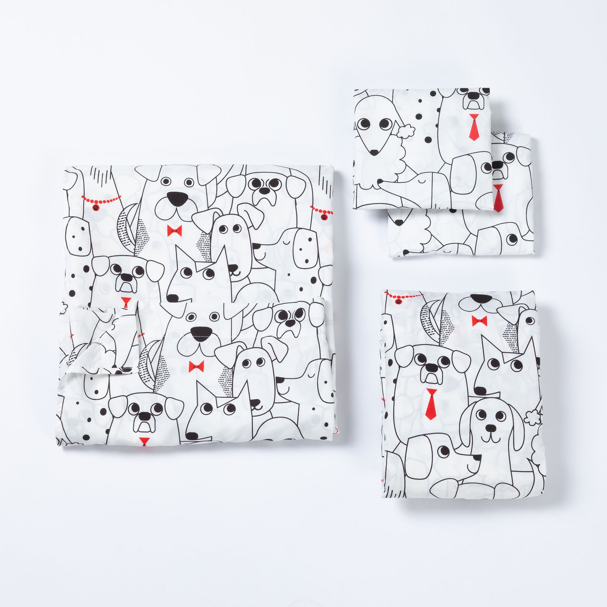 A set of folded sheets, including a larger square piece and three smaller rectangular pieces, all printed with a playful black-and-white dog pattern and small red bow tie and tie accents, arranged on a clean white background.