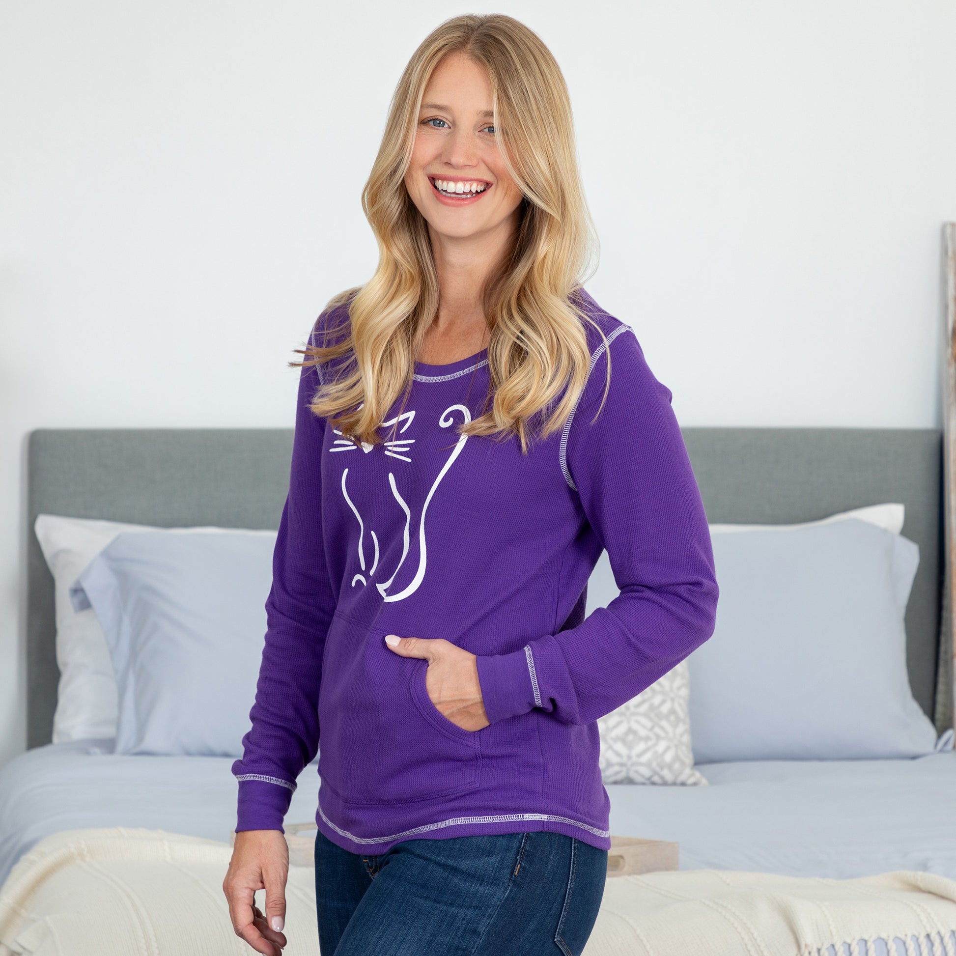 Woman smiling in a bedroom wearing a purple long sleeve thermal top with a cat silhouette on the front.