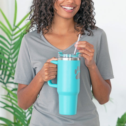 A person holding a teal 40 oz tumbler with straw, with a paw print on the front, standing in front of a white wall and greenery.