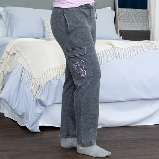 Gray sweatpants with a pink cat design, worn while standing beside a bed.