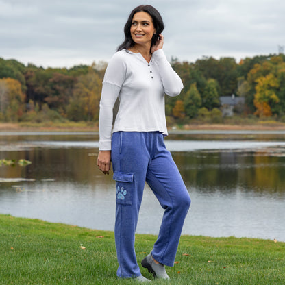 A woman stands on a grassy lakeside in autumn, wearing a white ribbed long-sleeve henley top and blue cargo-style pants with a blue paw print graphic on the thigh.