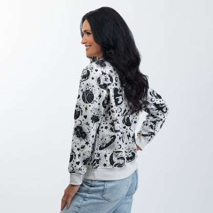 A woman wearing a light gray crewneck sweatshirt with a black celestial pattern of crescent moons, and stars, paired with light blue jeans, photographed against a light gray studio background.