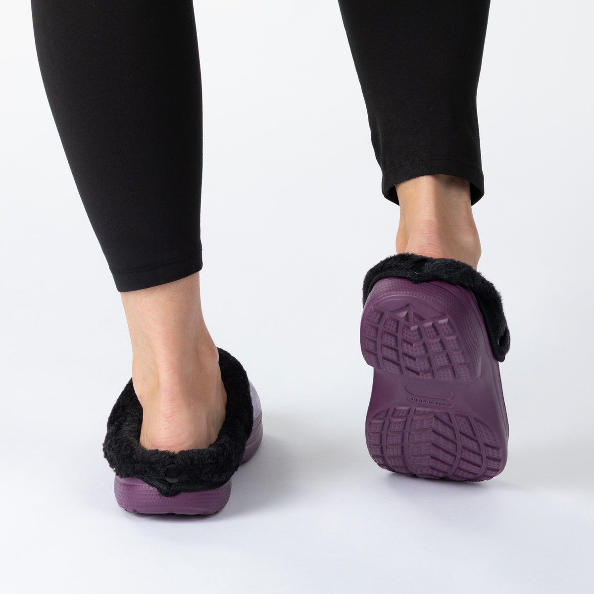 A person wearing black leggings and purple slip-on slippers with black faux-fur trim, shown from the back against a clean white studio background. One slipper is shown from the side/back and the other from the back.