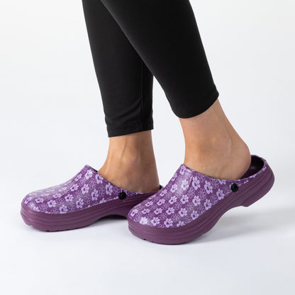A person wearing black leggings and purple paw print clogs with a small black button, shown from the calves down on a clean white studio background.