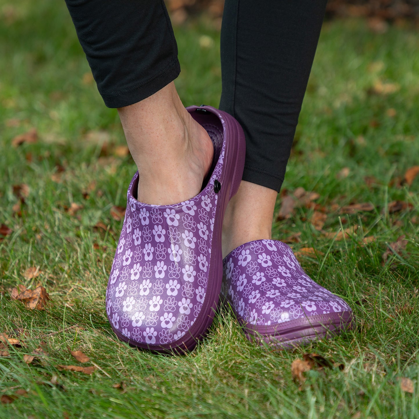 A person wearing purple slip-on clogs with a light paw print pattern, standing on green grass with scattered brown leaves.