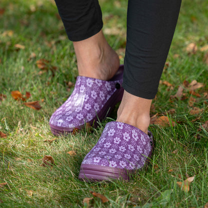 Purple clogs with a white floral pattern worn by a person standing on grass with scattered autumn leaves.