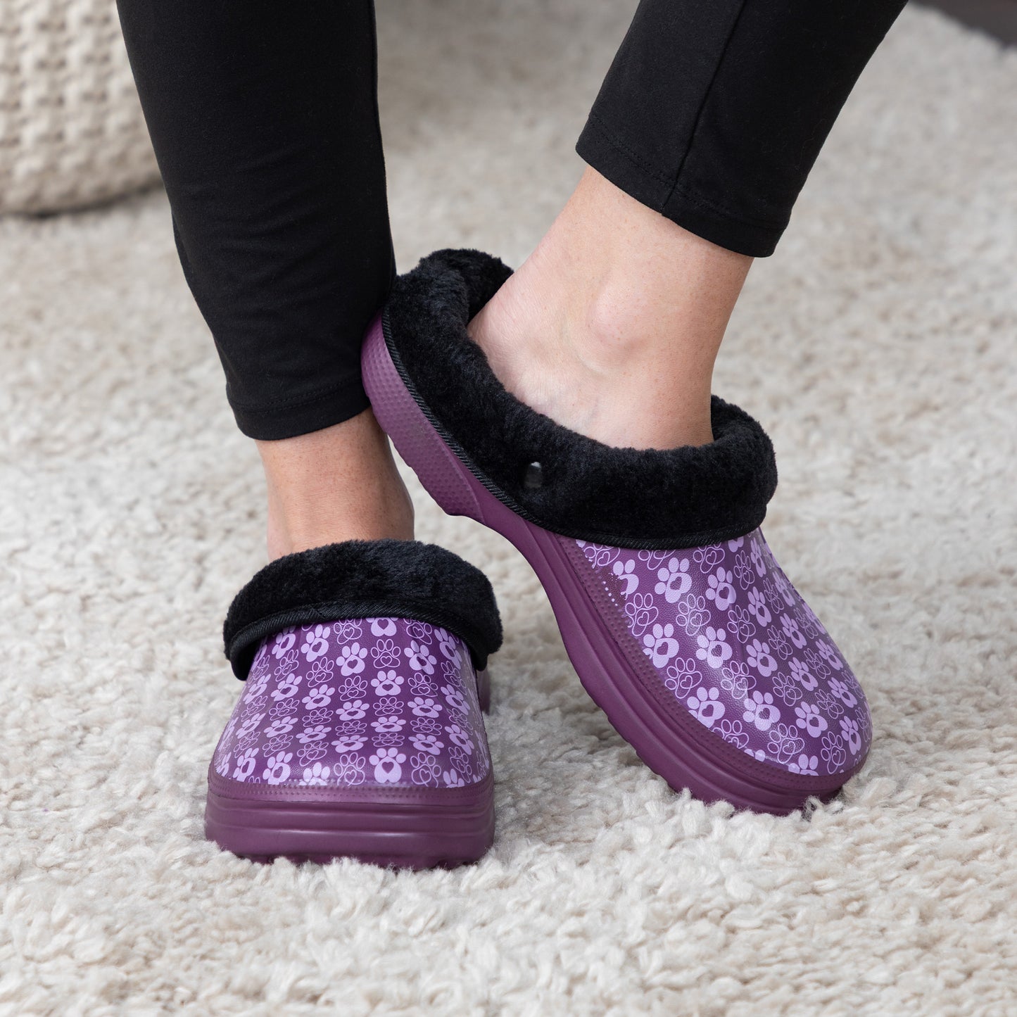 A person wearing purple slip-on clogs with a paw print pattern, black faux-fur trim, and thick purple soles, standing on a cream-colored carpet.