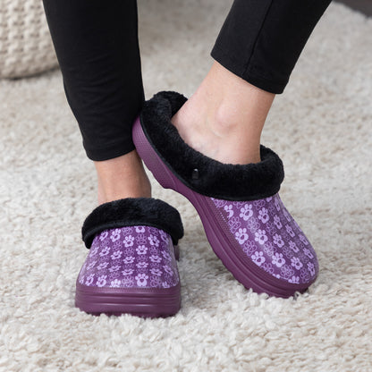 A person wearing purple slip-on clogs with a paw print pattern, black faux-fur trim, and thick purple soles, standing on a cream-colored carpet.