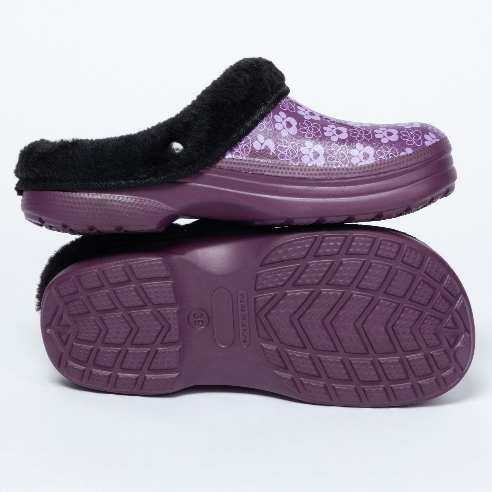 A pair of women's clogs shown on a white background, with one shoe standing upright to display the top and the other lying flat to show the sole. The top features a purple and white paw print pattern with a black faux-fur trim, while the sole is a solid dark purple with a textured tread pattern.