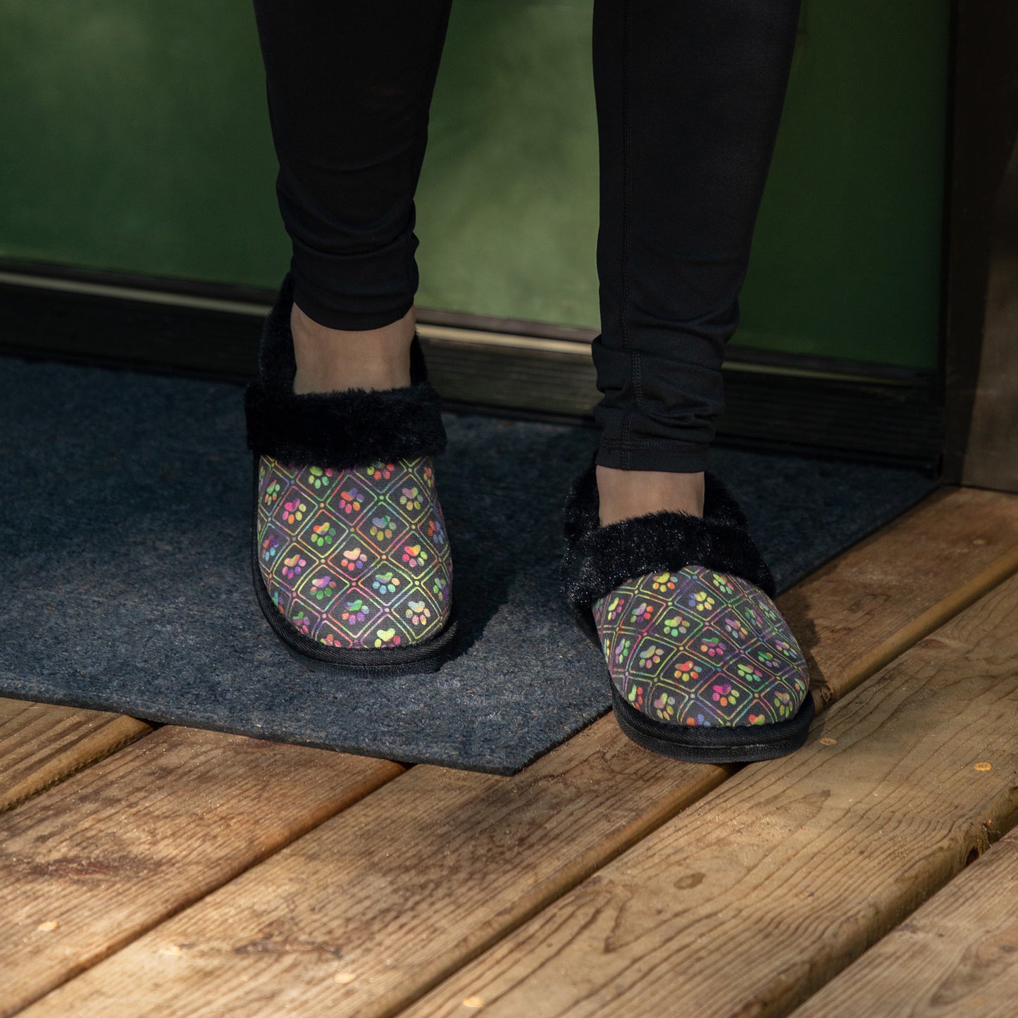 Person wearing black faux fur lined paw printed slipper shoes on wood deck.