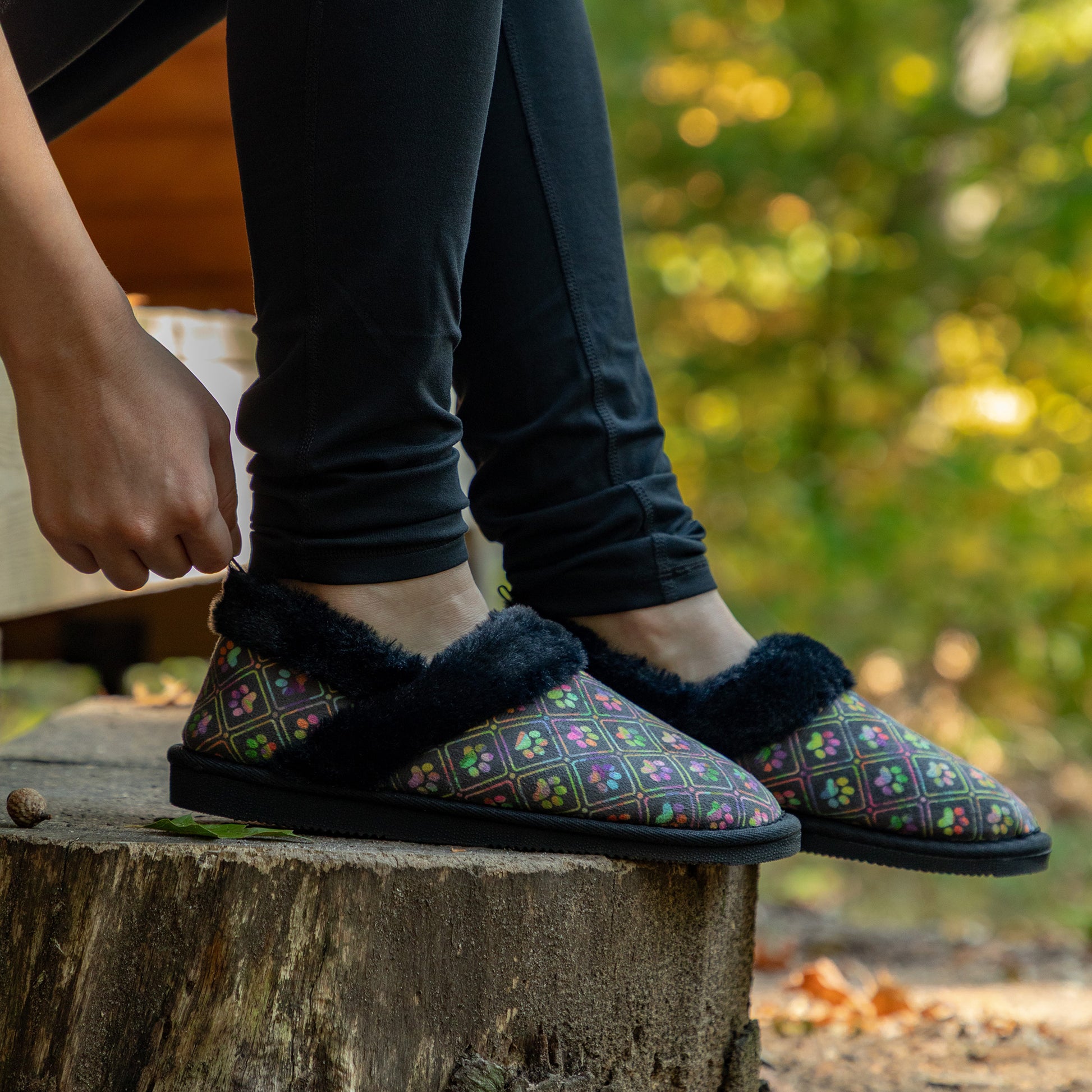 Person wearing black faux fur lined paw printed slipper shoes in outdoor wooded setting.