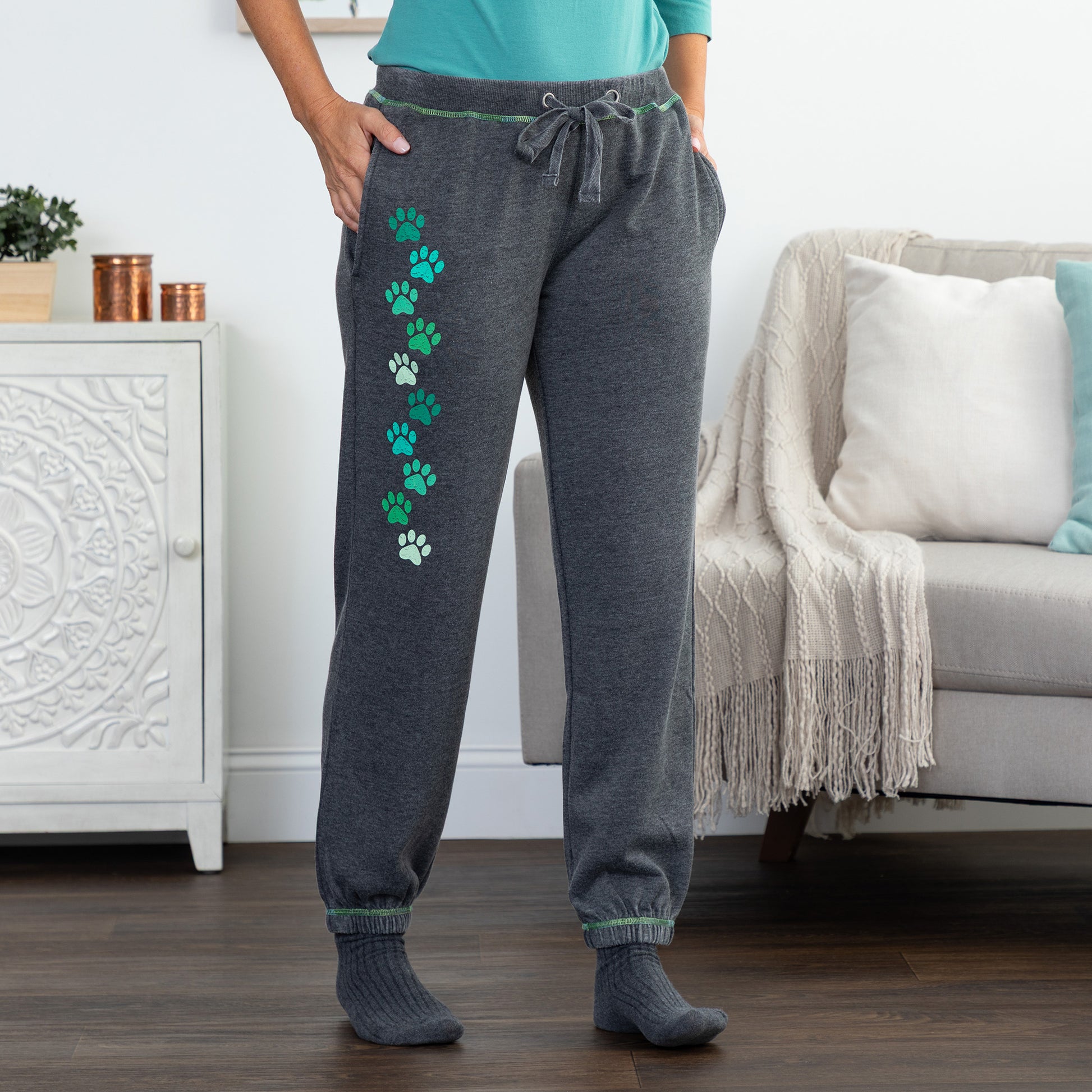 A person wearing dark gray jogger sweatpants with a vertical row of green and white paw prints down the left leg, standing in a living room with a sofa and cabinet in the background.