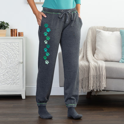 A person wearing dark gray jogger sweatpants with a vertical row of green and white paw prints down the left leg, standing in a living room with a sofa and cabinet in the background.