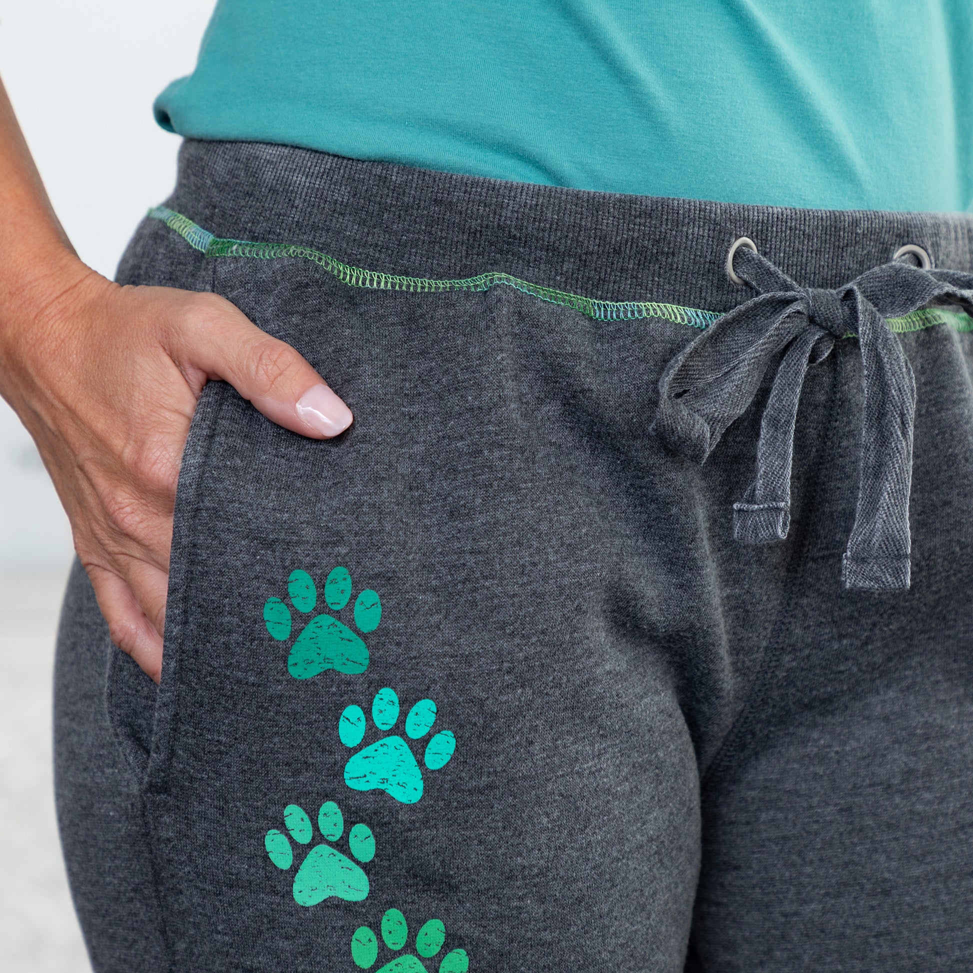 Close-up of a person wearing dark heather gray jogger sweatpants with a green drawstring and green paw print graphics, paired with a teal top.