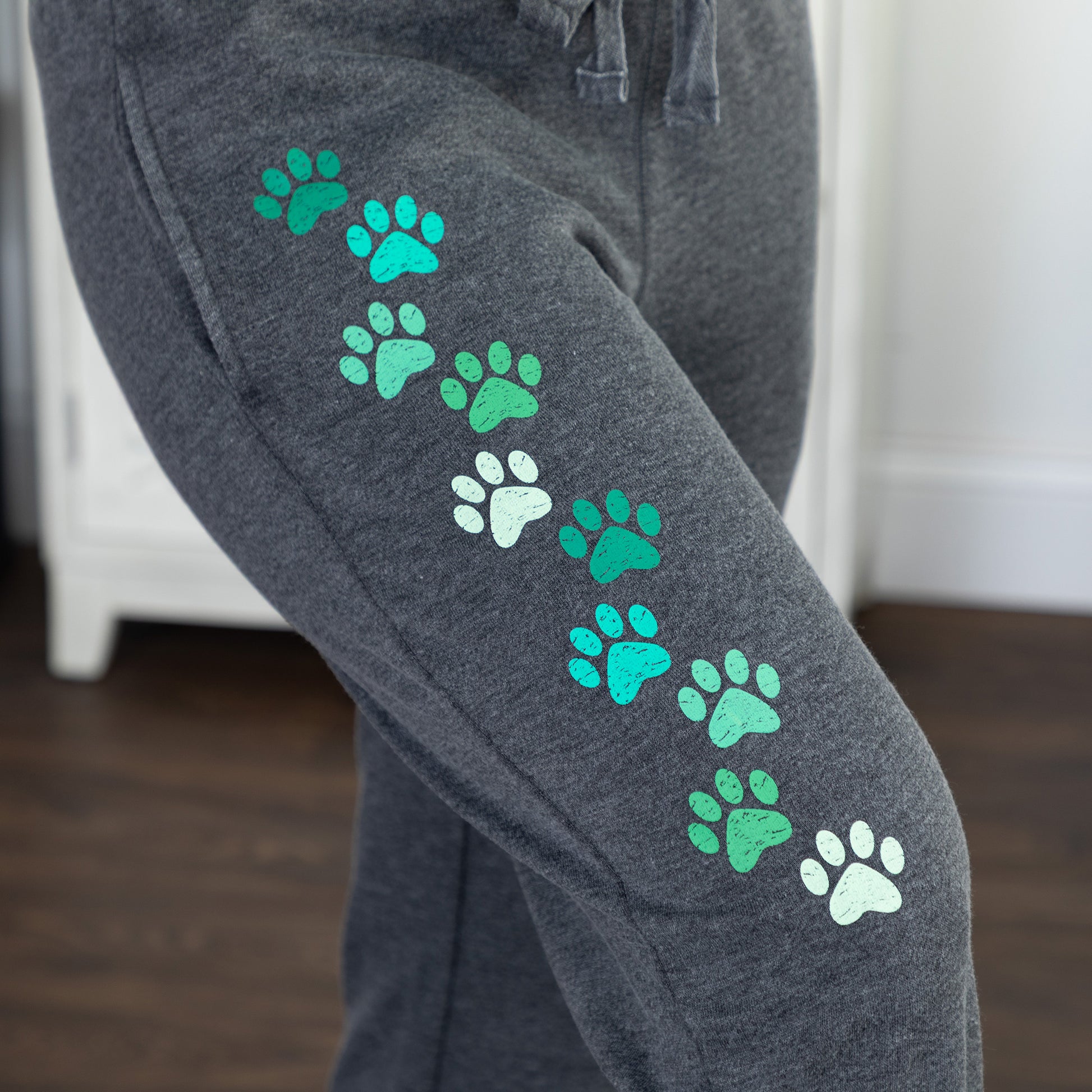 Close-up of a person wearing dark heather gray sweatpants with a diagonal pattern of green and mint paw prints on the upper leg, standing in an indoor room with a wooden floor and white furniture in the background.