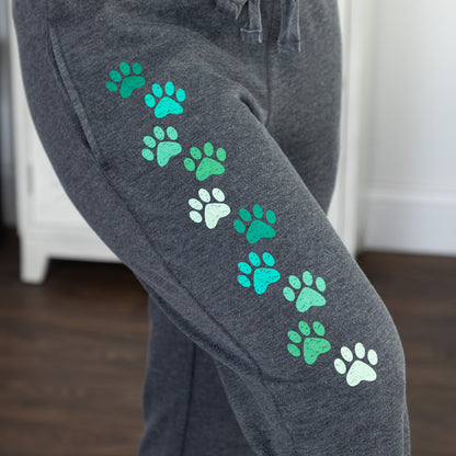 Close-up of a person wearing dark heather gray sweatpants with a diagonal pattern of green and mint paw prints on the upper leg, standing in an indoor room with a wooden floor and white furniture in the background.