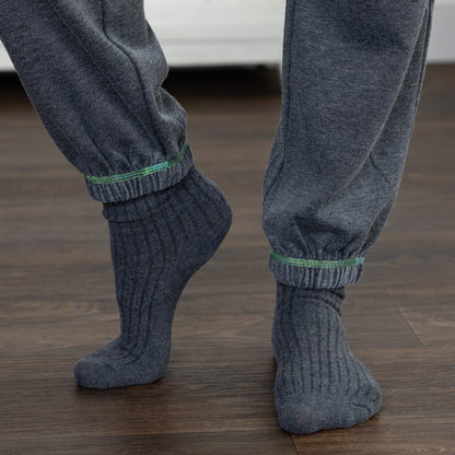 Close-up of a person's feet wearing dark gray ribbed socks with a thin green stripe detail at the ribbed cuffs, paired with matching dark gray jogger pants on a dark wood floor.