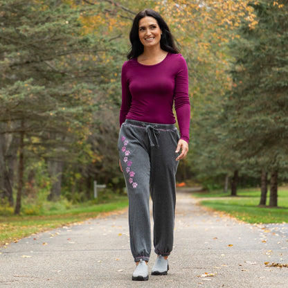 A woman walking on a paved path outdoors, wearing a fitted magenta long-sleeve top and dark gray jogger sweatpants with pink paw print embroidery on the left leg.