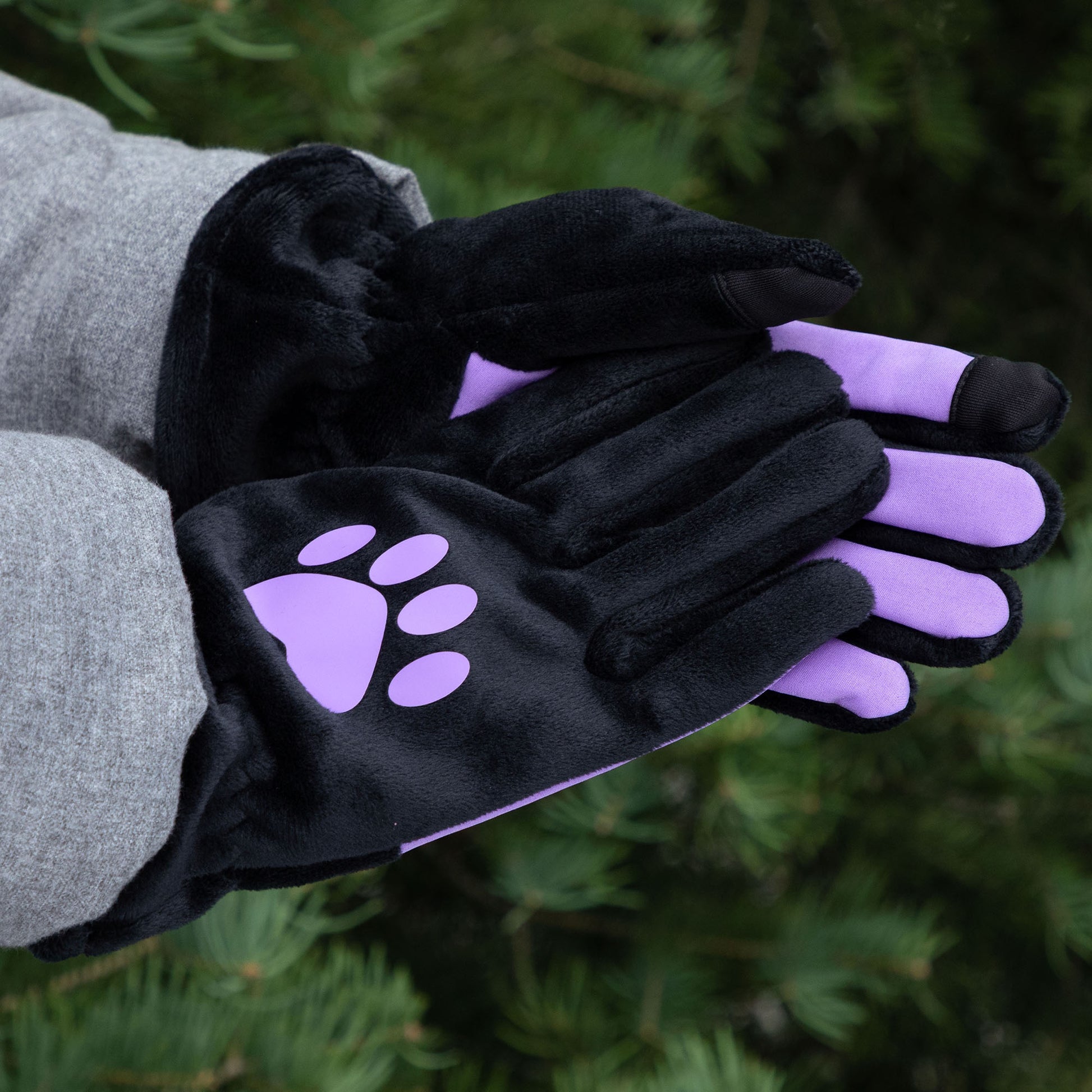 A person wearing black fleece gloves with purple paw prints against a green outdoor background.