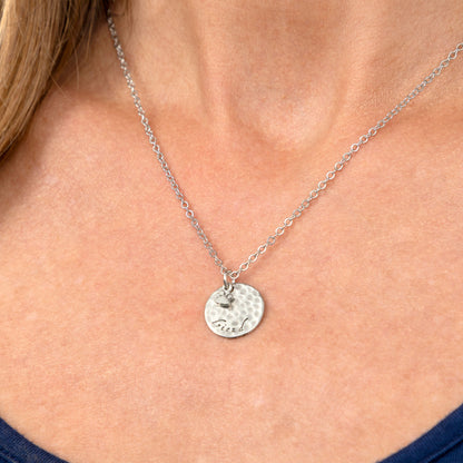 A close-up of a person's neck and chest wearing a silver chain necklace with a hammered round pendant engraved with 'friend'.