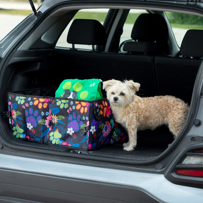 A foldable car trunk organizer with a paw print design is placed in the open trunk of a car. A small dog is standing beside it.