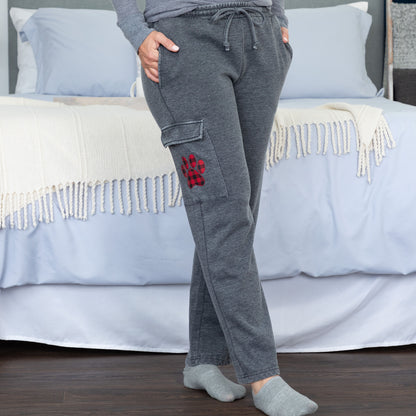 A person wearing dark gray drawstring sweatpants with cargo pockets, standing in front of a bed with white bedding and a cream fringed throw blanket. The pants have a red and black buffalo plaid pattern on the cargo pocket.