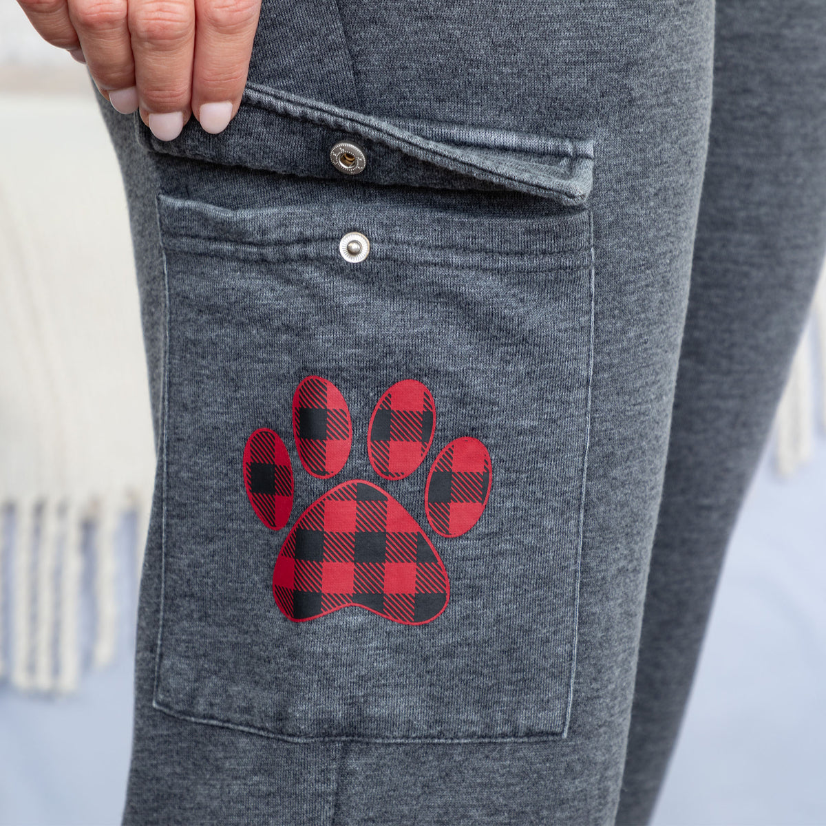 Close-up of a person wearing dark heather gray pants with a red and black buffalo plaid paw print patch on the pocket.
